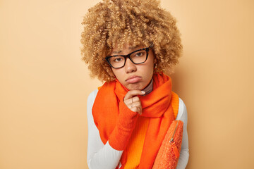 Portrait of distressed upset female model keeps hand on chin feels frustrated wears spectacles orange scarf around neck carries small bag under arm looks sadly at camera isolated over beige wall