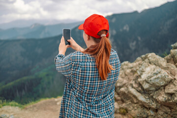 Fototapeta na wymiar Back view of young beautiful hiker woman taking selfie portrait on the top of mountain, using her smartphone - Hiking and climbing cliff 