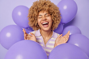Happy beautiful woman with curly hair keeps palms raised up laughs joyfully dressed in striped jumper glad to celebrate special occasion surrounded by inflated balloons poses against purple background