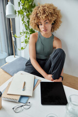 View from overhead of curly haired young woman poses in modern quiet workspace uses tablet...