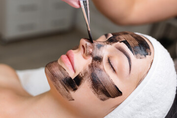cosmetologist applying black mask on pretty woman face wearing black gloves, gorgeous woman in spa having facial procedures