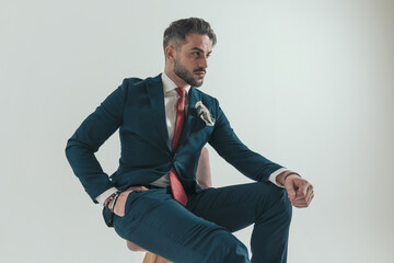 confident fashion guy in suit with hand in pocket sitting and looking to side