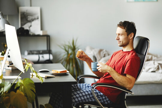 Young Man Eating Sandwich And Drinking Coffee While Sitting On Chair In Front Of Computer And Watching Video