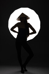 Disco, nightlife, retro, fashion concept. Black silhouette of slim and sexy woman with cone-shaped Asian cane hat in front of bright illuminated white light with copy space. Abstract studio portrait