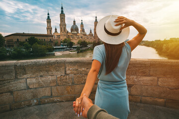 Follow me. Tourist woman holding man's hand and walking in Zaragoza city. The Cathedral-Basilica of Our Lady of the Pillar on sunset. Couple on vacation