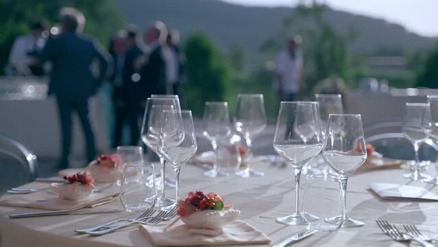 Exit ceremony in luxury restaurant, crystal goblets shining and reflecting in sunset on beautifully decorated table. Drinking glasses, silverware and flowers bouquets as a part of ceremony decoration