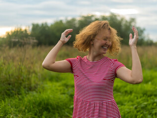 a girl in a red summer dress with red hair flowing from the wind against a background of a green meadow and a sky with clouds