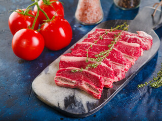On a dark blue background, a cutting board with raw meat steaks with a sprig of rosemary, appetizing ripe tomatoes and spices. Close-up. Cooking meat steaks according to the classic recipe.