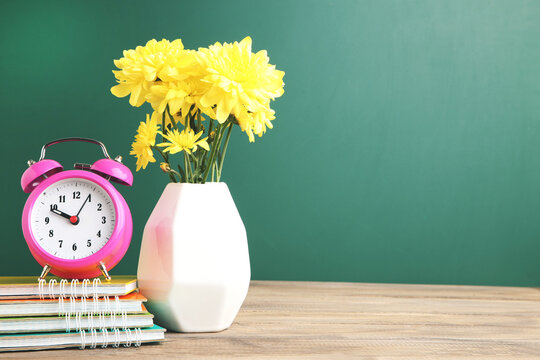 Chrysanthemums in vase, alarm clock and colorful notebooks on school chalkboard background with copy space. Concept teachers day