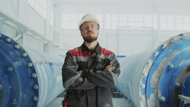 Medium portrait of confident young male plant worker in uniform posing with hands folded against two grinding machines processing marble aggregate used in construction