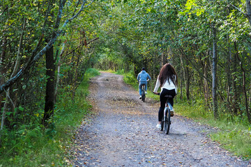 Fototapeta premium Young man and woman on bicycles on narrow bike path through summer trees in the forest. Bikers on a ride in a park