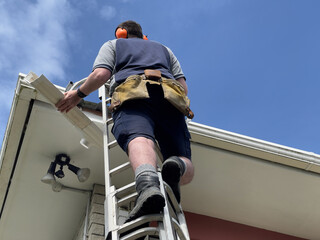 Young caucasian man in his 30's wearing earmuffs standing on a portable scaffold ladder working to replace a section of plastic guttering on a house in New Zealand © Harriet