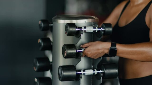 Sport training with equipment. Close up shot of unrecognizable black woman athlete taking dumbbells from rack at gym