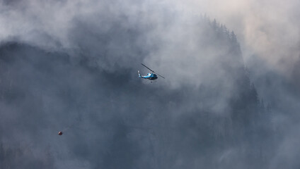 Naklejka premium Wildfire Service Helicopter flying over BC Forest Fire and Smoke on the mountain near Hope during a hot sunny summer day. British Columbia, Canada. Natural Disaster