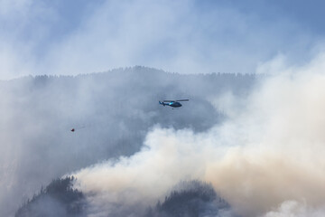 Naklejka na ściany i meble Wildfire Service Helicopter flying over BC Forest Fire and Smoke on the mountain near Hope during a hot sunny summer day. British Columbia, Canada. Natural Disaster