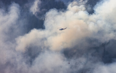 Fototapeta premium Wildfire Service Helicopter flying over BC Forest Fire and Smoke on the mountain near Hope during a hot sunny summer day. British Columbia, Canada. Natural Disaster
