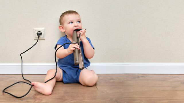 Child boy chewing on the electric wire of a curling iron, copy space. Toddler baby plays with hot curling tongs at the electrical outlet. Kid age one year