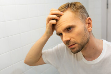 Middle aged caucasian white man with a short beard looks at his hair in the mirror in the bathroom and worried about balding. The concept of the problem of male hair loss, early baldness and alopecia