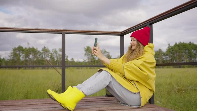 Pretty woman in a red hat and yellow poncho raincoat takes a selfie while sitting on a wooden platform in the forest