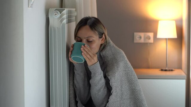 Young woman covered with warm grey blanket holds mug of hot beverage. Female gets cold and feels unwell sitting on cold floor. Blonde lady snuggling up to central heater tries to warm up in evening
