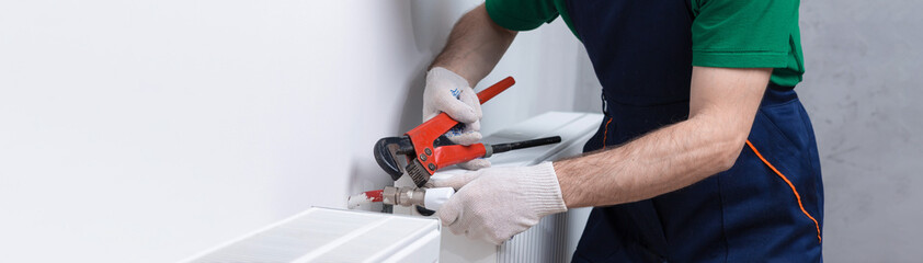A male plumber installs a radiator in the heating system of an apartment. Guy in overalls and a gas...