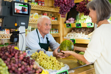 Senior salesman standing at counter in greengrocer and serving woman customer who purchasing fresh...