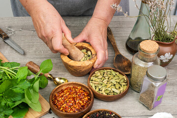 Woman's hands grind cardamom in a wooden mortar. Kitchen gray table with various spices.