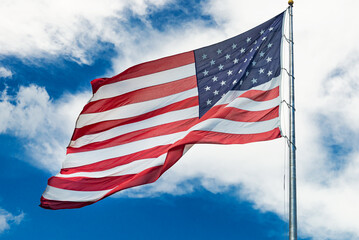 Obraz premium Large American flag waving in the wind against a cloudy blue sky. Low angle view of stars and stripes on american flag against blue sky