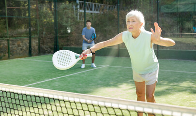 Fototapeta premium Portrait of active emotional elderly woman playing padel tennis on open court in summer, swinging racket to return ball over net ..