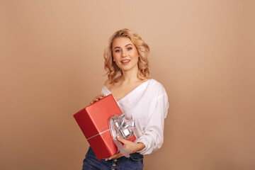 A smiling blonde woman in a white blouse holding a red gift box isolated on a beige background. Copy space.