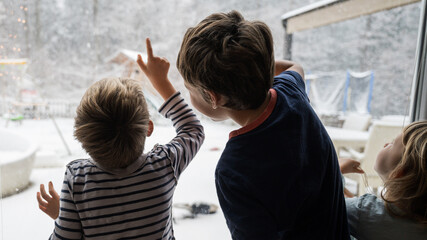 Three kids, brothers and sister, looking out the window pointing to beautiful winter nature