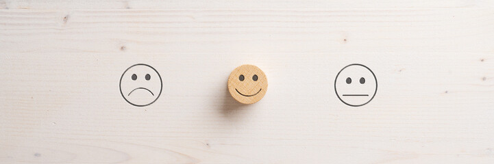 Sad and neutral emoji face symbols cut into wooden background and a smiling one in the middle