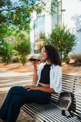 Shot of black woman sending a voice message to friends while she is sitting on a bench in the park