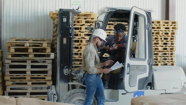 Warehouse worker talking about goods transportation to African American female colleague sitting in truck lift