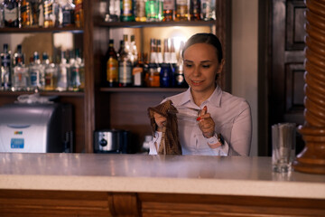 Woman bartender rubs the glass in cafe. Girl barista in casual clothes smiling and using rag wiping glass at counter in bar
