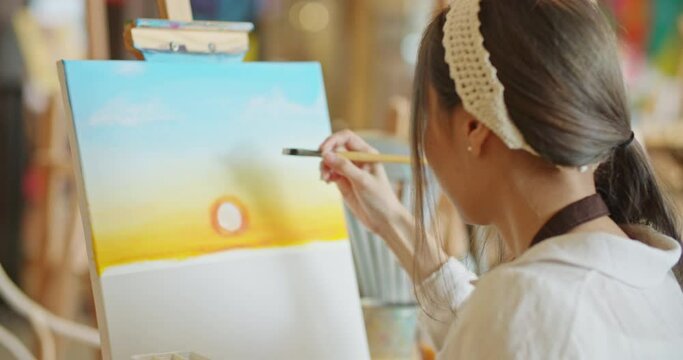 Back view of Attractive beautiful asian stylish female artist painting picture in art studio. Young woman painter draws with paint brush and oil colors creating artwork sunset landscape on canvas.