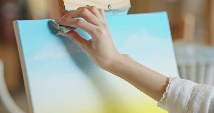 Close-up Shot of female artist hand using a sponge applying paint on canvas drawing painting picture in art studio. Young woman painter creating artwork sunset landscape. imagination and creativity.