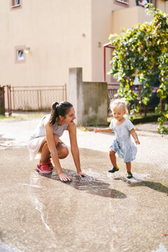 Smiling Mom Crouched Over A Puddle On Which A Little Girl Runs. High Quality Photo