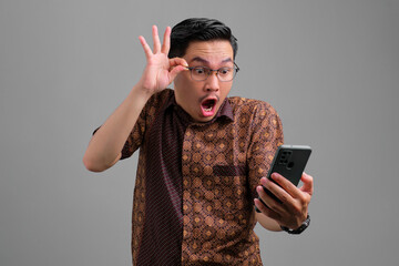 Shocked young Asian man wearing batik shirt looking at smartphone with stunned facial expression isolated on grey background, touching glasses