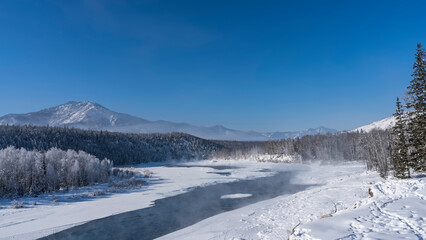 Fototapeta na wymiar An ice-free river in a snow-covered valley. Steam rises above the water. Footprints in snowdrifts. The trees in the forest are covered with hoarfrost. A mountain against a clear blue sky. Copy space