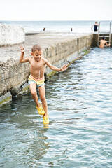 Fototapeta premium Small boy jumps from stone pier to sea against people. Moment of excited child jumping and touching water. Small kid looks like magician