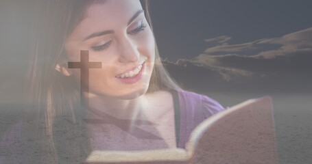 Multiple exposure of smiling caucasian young woman reading bible and silhouette cross against sky