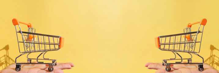 On a woman's hand is a small empty metal trolley from a supermarket. Banner. The object is on a yellow background. Horizontal photo. trading concept. Buyer. Shopping concept. Close-up. Copy space.