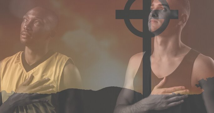 Caucasian and african american mid adult men with hand on chest praying with cemetery cross on land