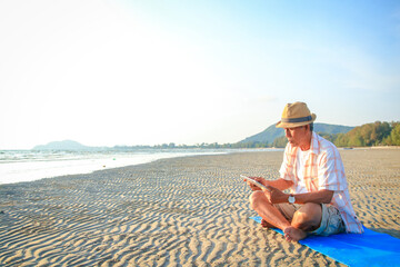 Asian elderly man sitting on the beach by the sea playing tablet He enjoyed his vacation. Tourism...