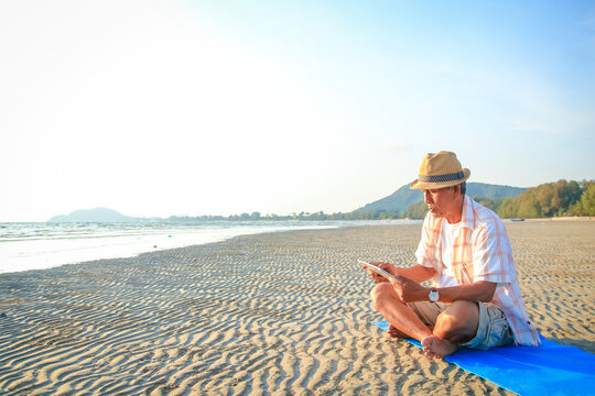 Asian elderly man sitting on the beach by the sea playing tablet He enjoyed his vacation. Tourism concept. Living in retirement. Elderly people use modern technology. Copy space