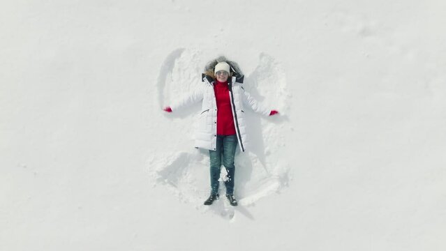 AERIAL 4K overhead view of young girl lying on fresh white snow, painting angel by moving her arms, legs up and down. Drone above happy positive smiling woman in winter coat and red Christmas sweater