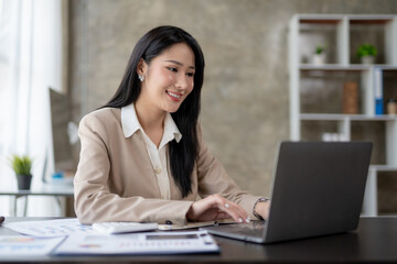 Beautiful Asian business woman using her laptop to work and enjoy working, taking note, reviewing assignment and smiling in the office.