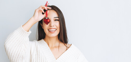 Young happy asian girl with long hair in white knitted cardigan with red heart christmas ball in...