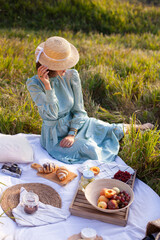 A girl in a long summer dress with short hair sitting on a white blanket with fruits and pastries...